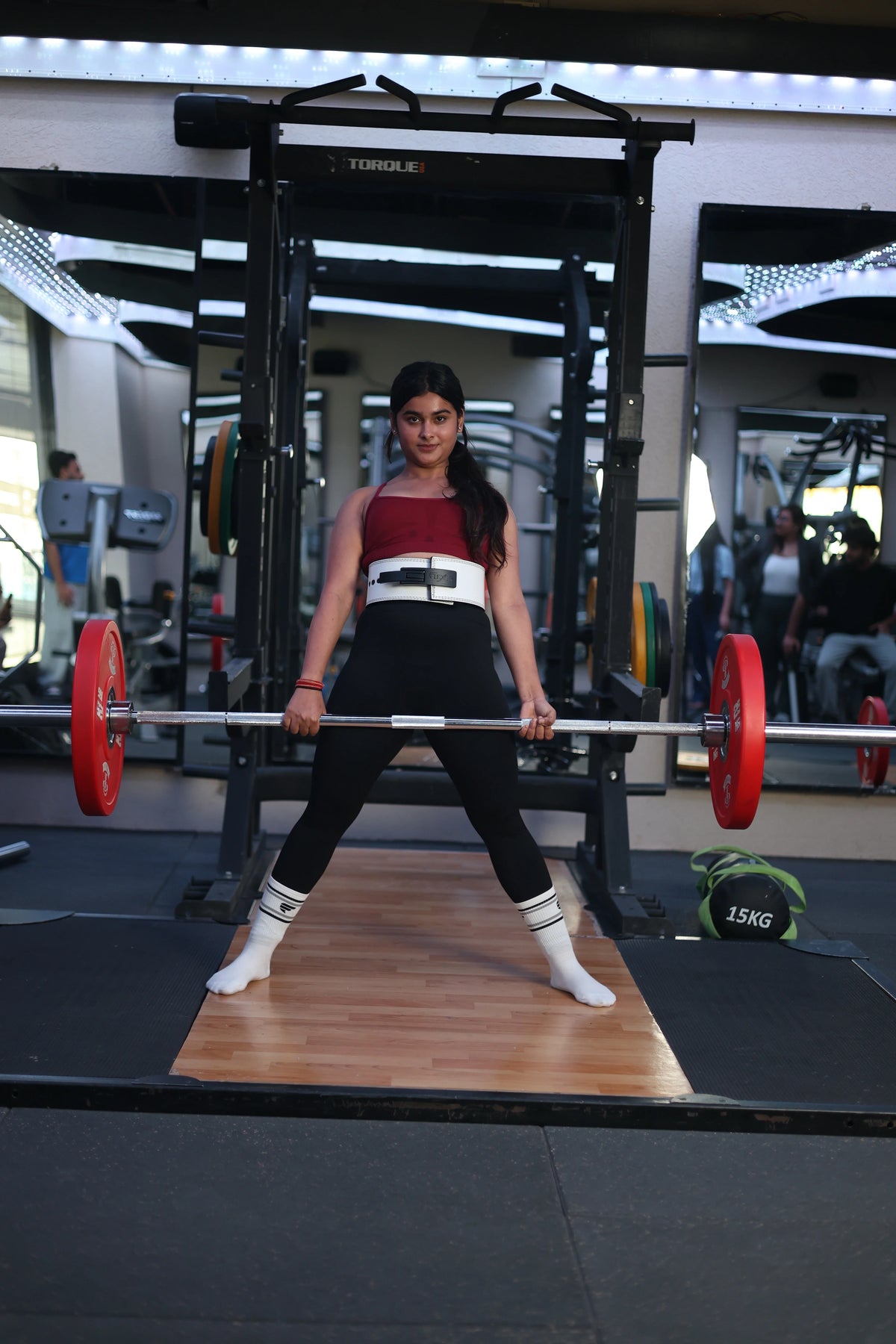 A woman stands in a wide sumo-style stance inside a gym power rack, holding a barbell at hip level during a deadlift. 