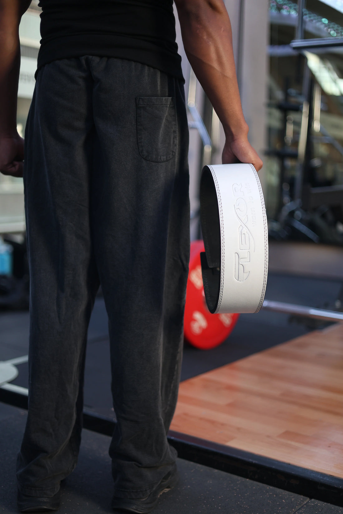 Rear view of a person standing in a gym, wearing dark trousers and a black shirt, holding a white leather weightlifting lever belt in their left hand